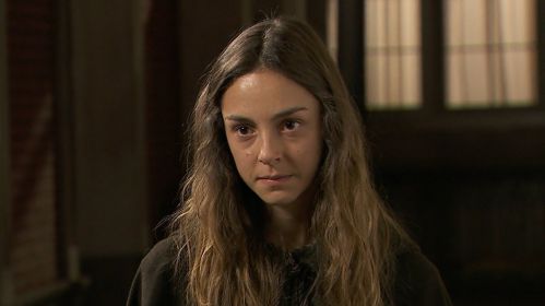 Olga spogląda z powagą i determinacją, a jej twarz zdradza emocje pełne napięcia. Długie, lekko potargane włosy opadają jej na ramiona, podkreślając surowy wygląd postaci. W tle widać przyciemnione wnętrze, które potęguje dramatyczny nastrój sceny z udziałem Olgi z serialu „Akacjowa 38”.