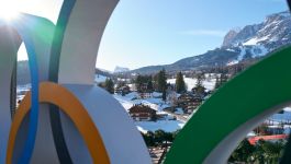 Widok zimowej scenerii w Cortina d’Ampezzo z górskimi szczytami i ośnieżonymi stokami w tle, drewnianymi domami w dolinie oraz fragmentem dużych olimpijskich pierścieni na pierwszym planie, symbolizujących nadchodzące Zimowe Igrzyska Olimpijskie Mediolan–Cortina 2026