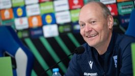 Trener Lecha Poznań, Niels Frederiksen, uśmiecha się podczas konferencji prasowej. Mężczyzna w średnim wieku, łysiejący, ubrany w niebieską kurtkę z logo klubu (lub sponsora), siedzi za mikrofonem. W tle widoczne są logotypy sponsorów na ściance konferencyjnej. Opis dotyczy zapowiedzi meczu Lecha Poznań z Rayo Vallecano w Lidze Konferencji.