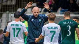 Trener Maciej Tokarczyk gratuluje piłkarzom Warty Poznań (fot. Warta Poznań)  Trener Warty Poznań Maciej Tokarczyk, ubrany w ciemną kurtkę i spodnie, stoi na boisku piłkarskim z uniesionymi obiema rękami, dając instrukcje trzem zawodnikom odwróconym do niego plecami. Dwóch z nich, w białych koszulkach z zielonymi numerami, to STEFANIAK z numerem 17 i WALUŚ z numerem 7. W tle widać fragment trybun z kibicami. Scena ilustruje rolę trenera w budowaniu drużyny i wprowadzaniu nowej filozofii, o czym Maciej Tokarczyk mówił w wywiadzie dotyczącym odrodzenia Warty Poznań