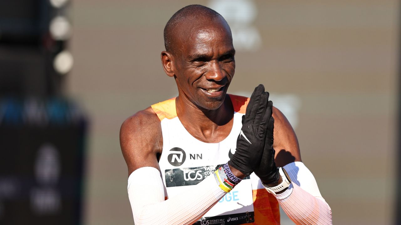 Na zdjęciu widzimy Eliuda Kipchoge, legendarnego kenijskiego maratończyka, uśmiechającego się i wykonującego gest podziękowania po przekroczeniu mety maratonu. Ubrany w charakterystyczny pomarańczowy strój startowy, z numerem startowym i rękawami kompresyjnymi, Kipchoge emanuje pewnością siebie i sportową elegancją. Fotograf uchwycił go w momencie celebracji, co doskonale podkreśla jego status jednej z największych gwiazd biegów długodystansowych na świecie.