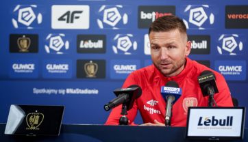 Daniel Myśliwiec, nowy trener Piasta Gliwice, w czerwonej koszulce, siedzi za stołem podczas konferencji prasowej, przemawiając do mikrofonów na tle tablicy z logo sponsorów, w tym 4F, Lebull i Gliwice. Po lewej stronie widać ekran z herbem Piasta Gliwice.